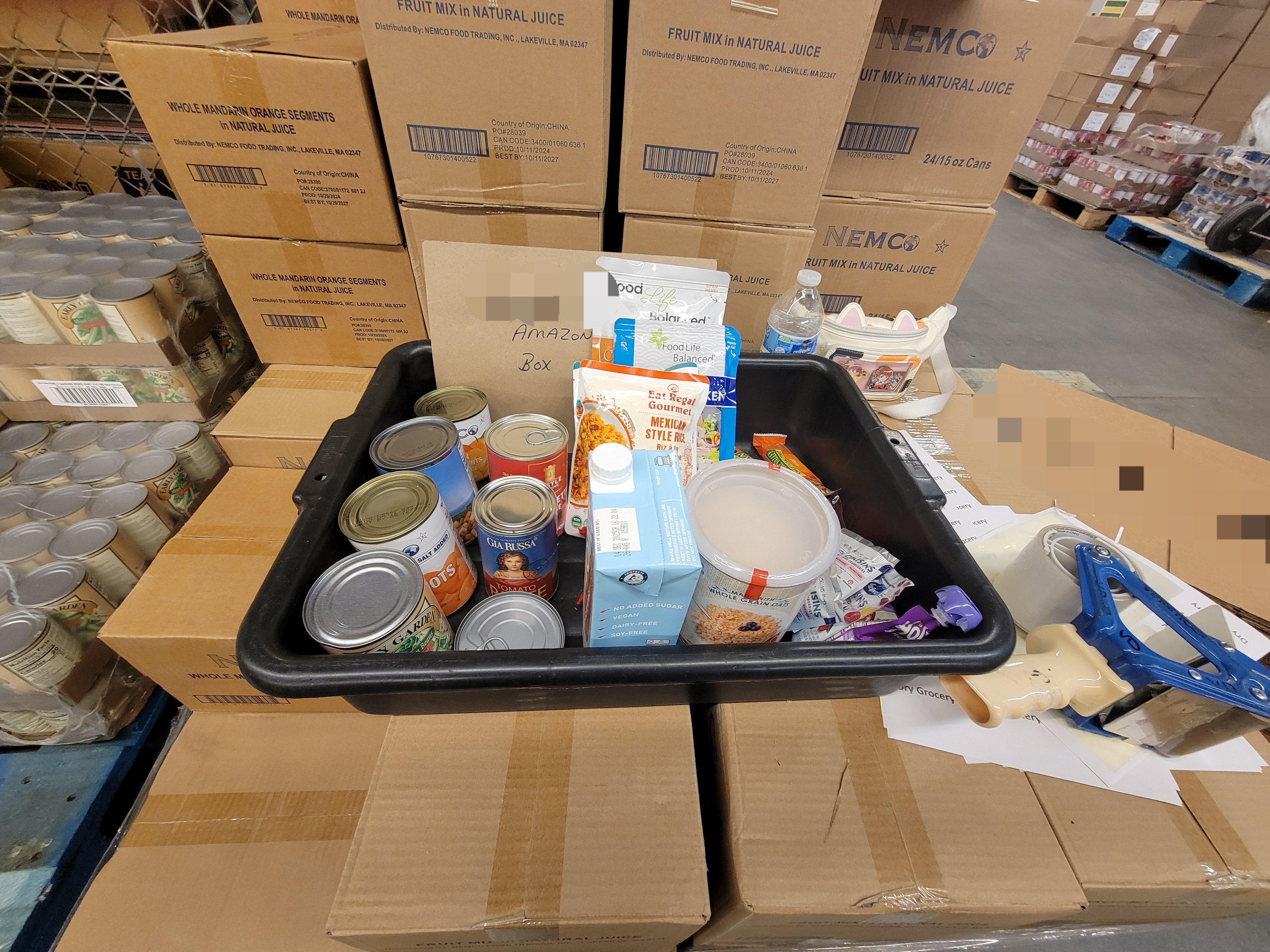 Tub of dry goods with a sign listing amazon box on top of cardboard boxes with tape gun and cat fanny next to it.